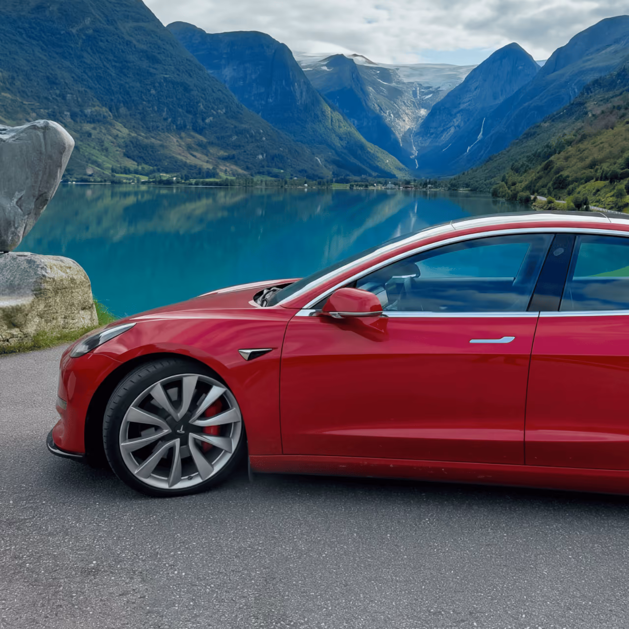 Red Tesla Model 3 by a fjord in front of green mountains.