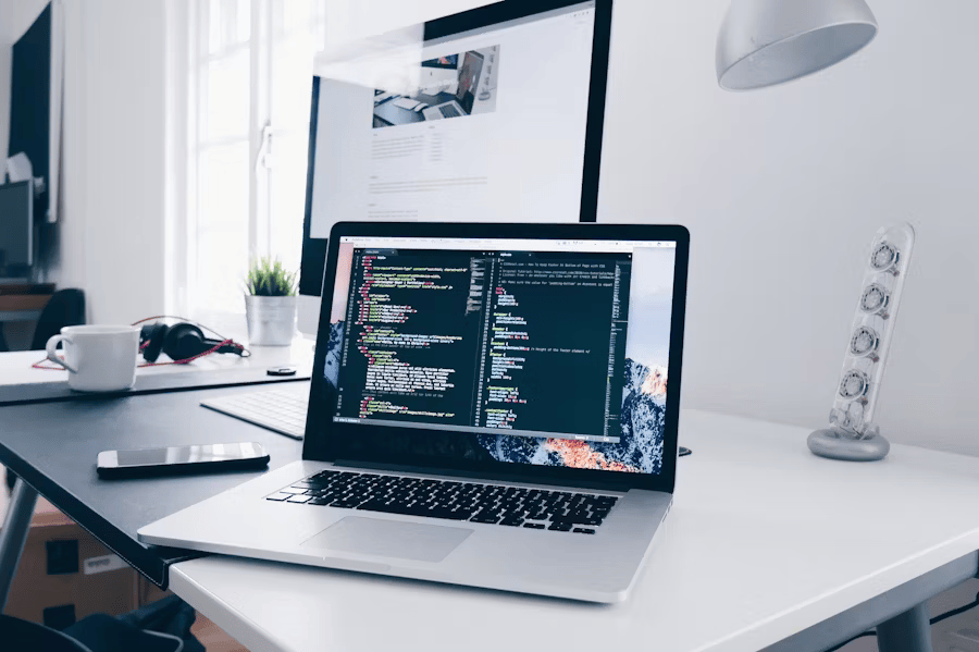 Ein moderner und heller Arbeitsplatz mit Fokus auf Webentwicklung. Im Vordergrund steht ein geöffneter Laptop, auf dessen Bildschirm Codezeilen zu sehen sind. Im Hintergrund befinden sich ein größerer Desktop-Monitor, ein Smartphone, eine weiße Tasse und Lautsprecher, was eine professionelle und technologieorientierte Arbeitsumgebung suggeriert.