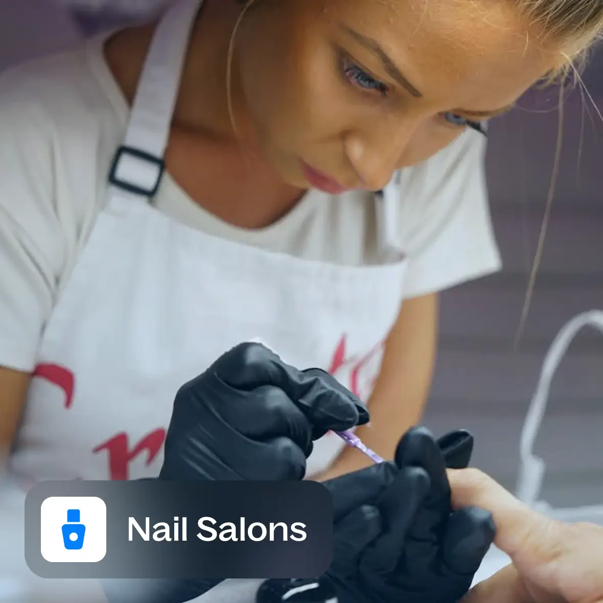 Woman wearing black gloves applying nail polish to a client's nails in a nail salon.