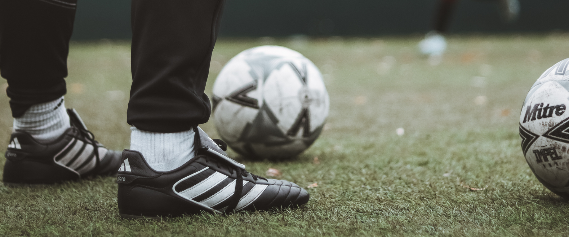 Close-up of a person wearing black Adidas soccer cleats and white socks standing on grass near two white and black soccer balls.