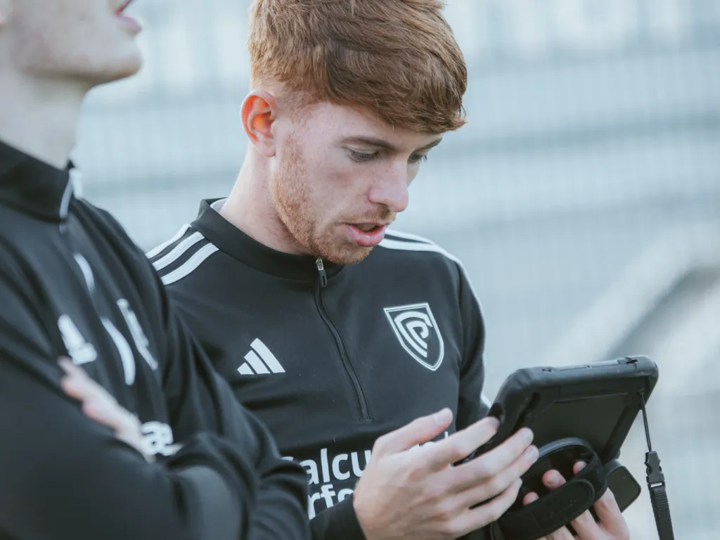 Young man in a black sports jacket looking intently at a tablet device held in his hands, with another person partially visible beside him.