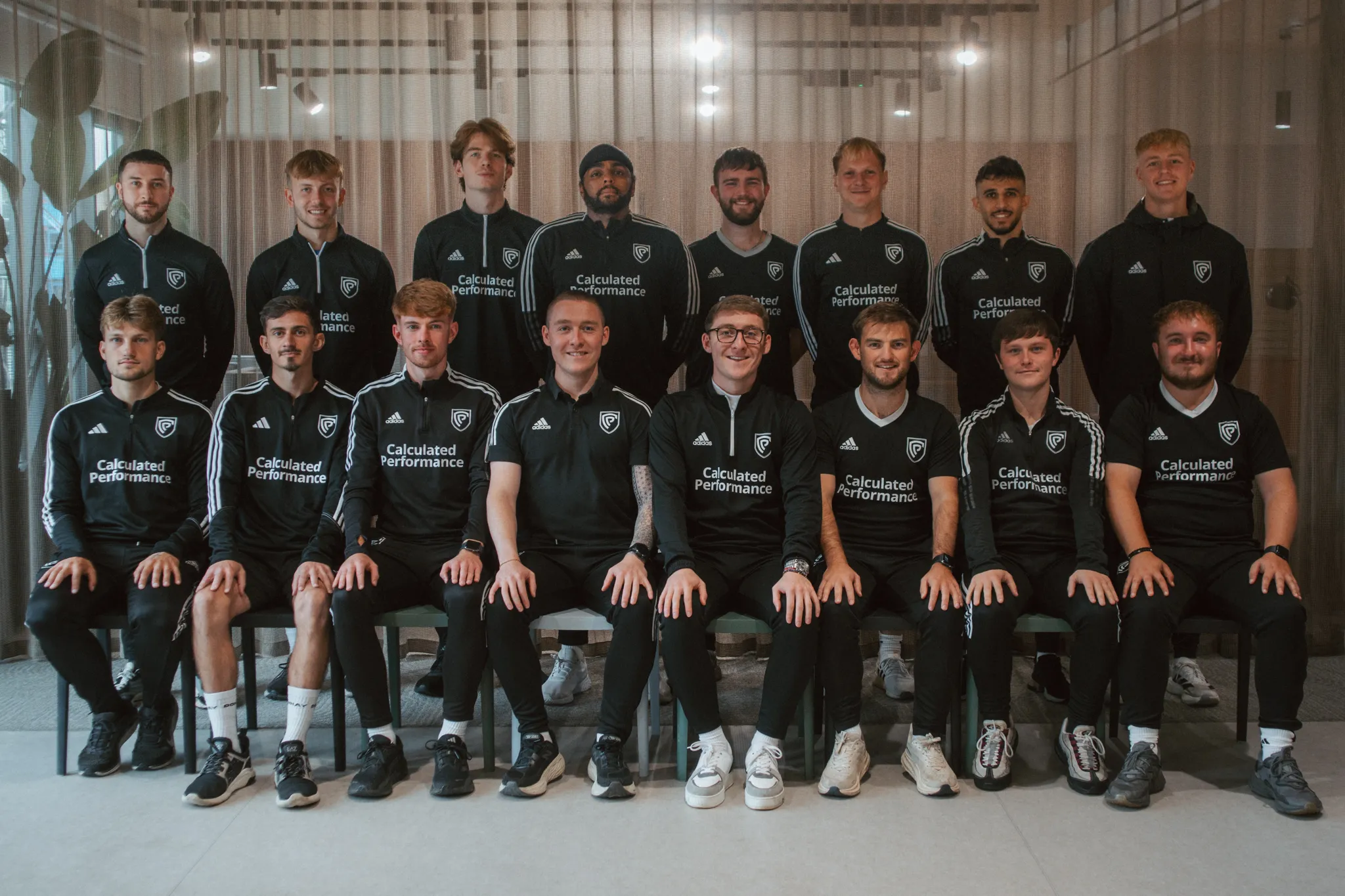 Group of fifteen men in black sportswear with Calculated Performance logo posing indoors in two rows, some sitting and some standing.