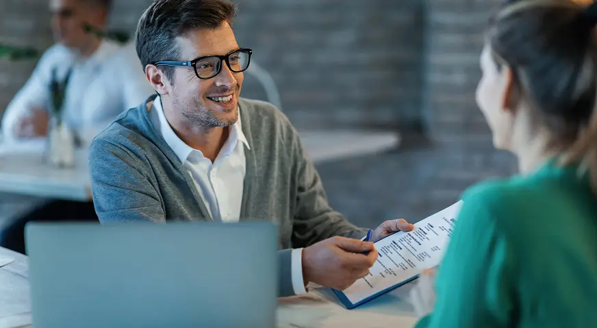 Man in glasses smiling and holding a clipboard, talking to a woman in a green sweater at a desk with a laptop.