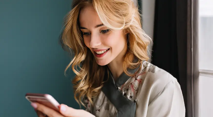 Smiling young woman with blonde hair looking at her smartphone near a window.