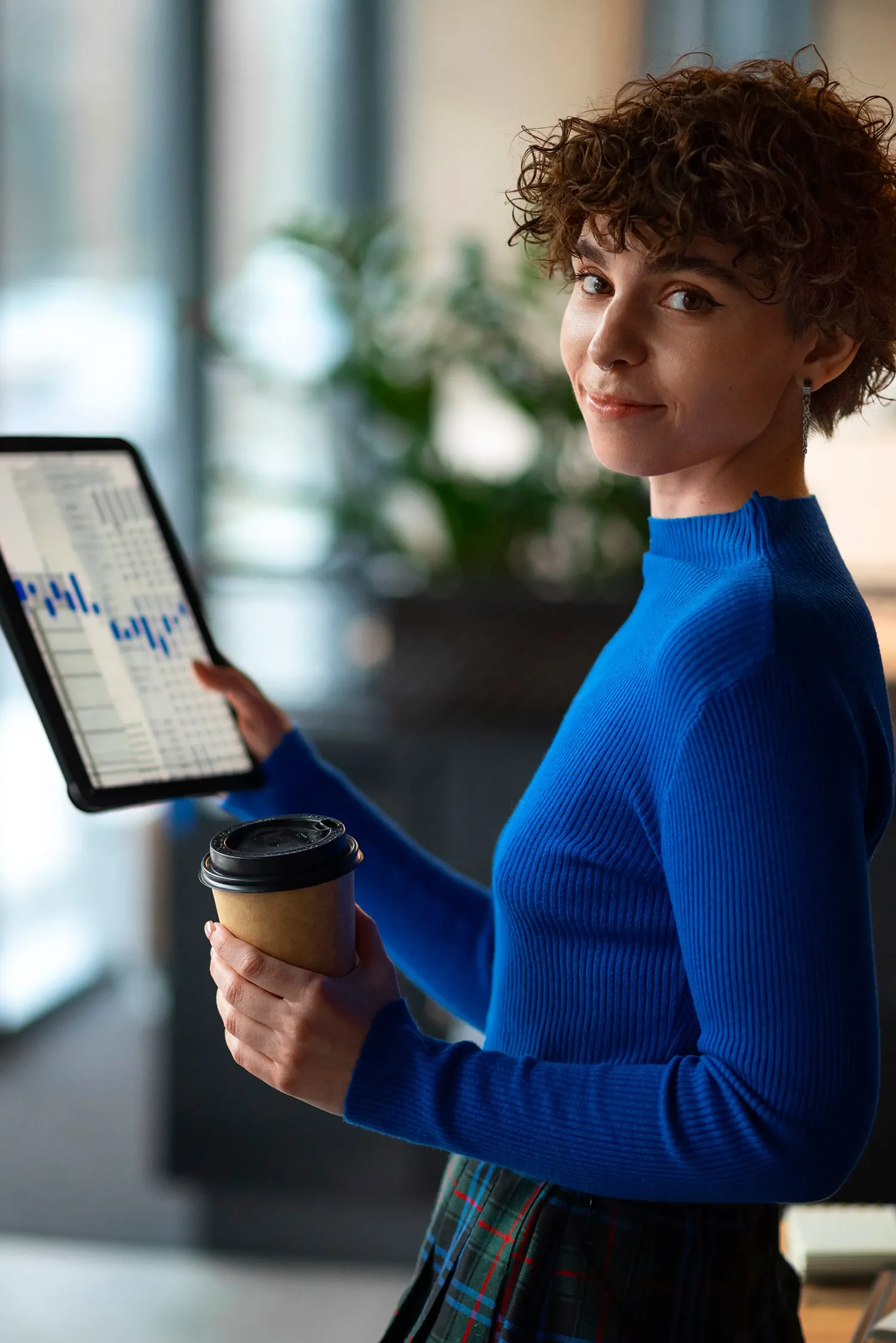 Young woman in a blue sweater holding a coffee cup and a tablet displaying a graph with blue bars in an office setting.