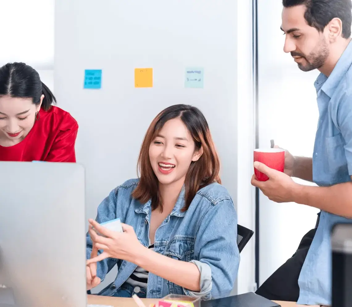Three colleagues collaborating in an office, one woman in a denim jacket smiling and holding a sticky note, another woman in red looking down, and a man holding a red cup.