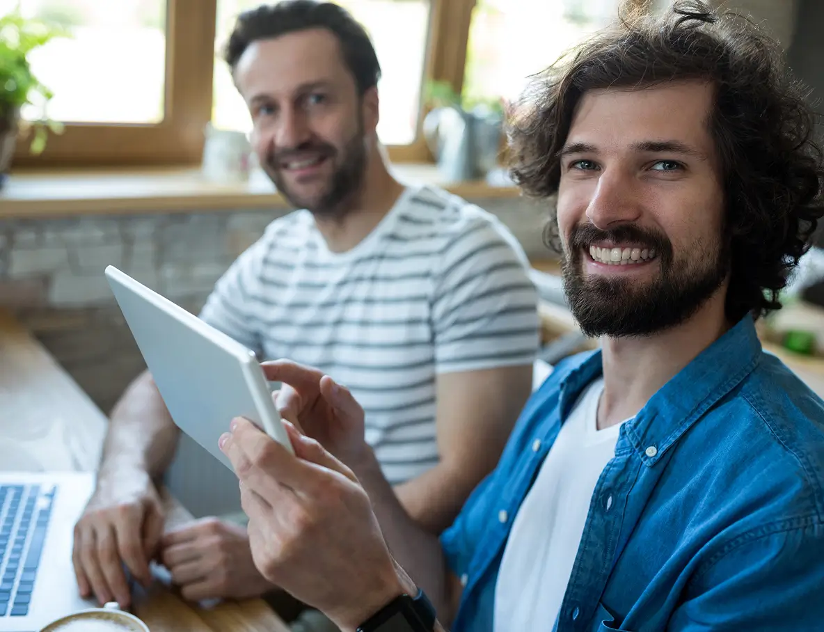 Two men smiling in a bright room, one holding a tablet and the other sitting near a laptop.