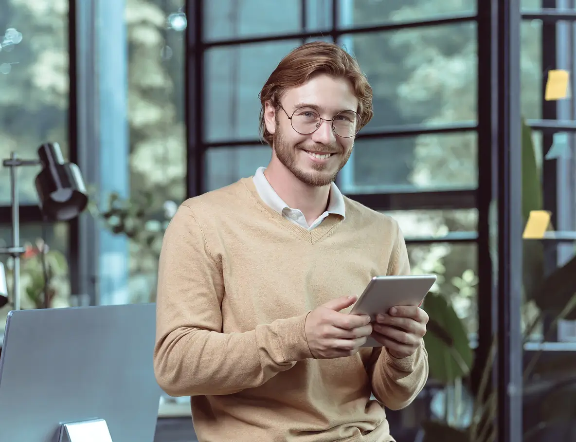 Smiling young man with glasses and light brown sweater holding a tablet in a modern office with large windows.