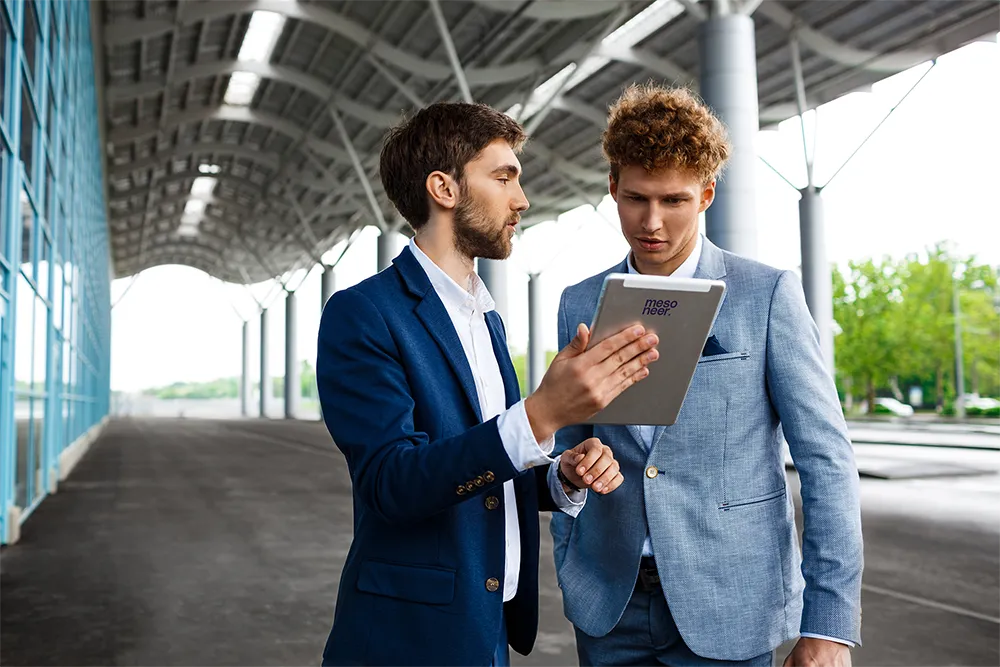 Two young businessmen in suits discussing info on a tablet under a modern outdoor canopy.
