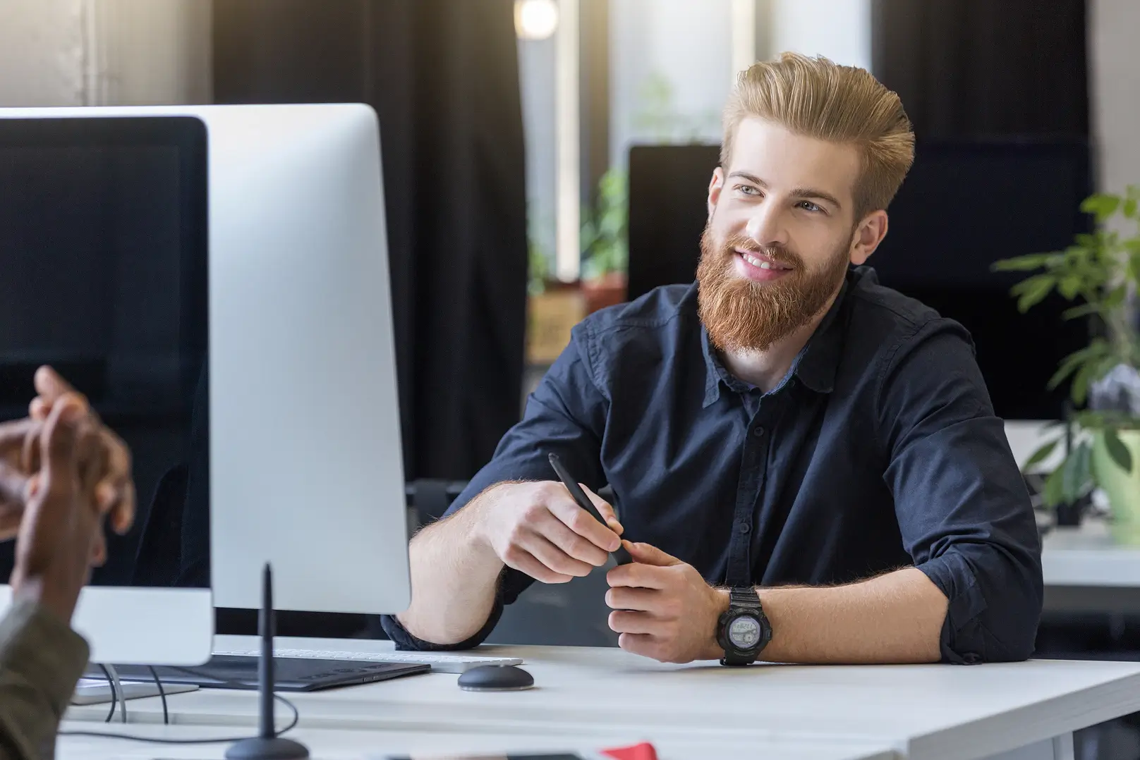 Smiling young man with a beard holding a pen and talking to a colleague in an office.