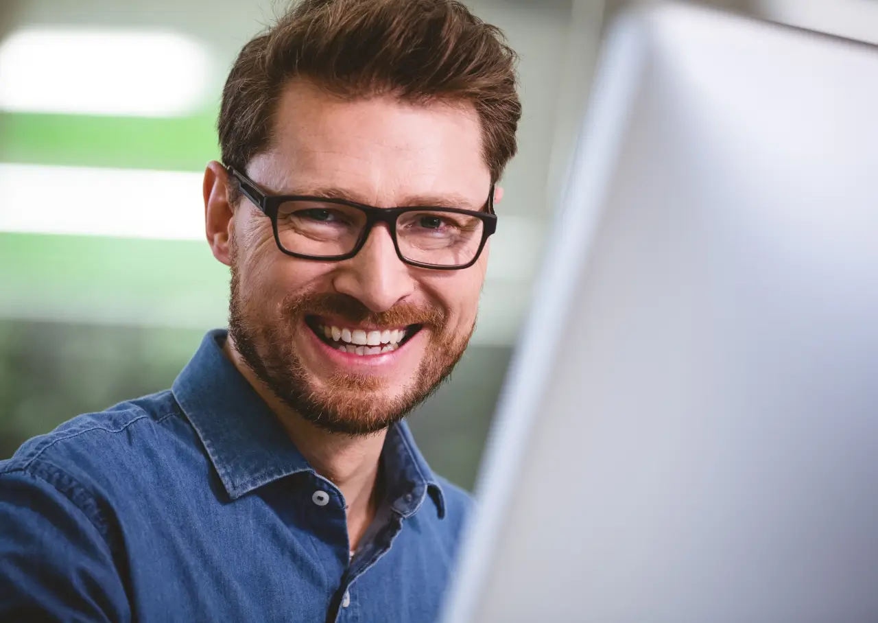 Smiling man with glasses and beard looking at a computer screen.