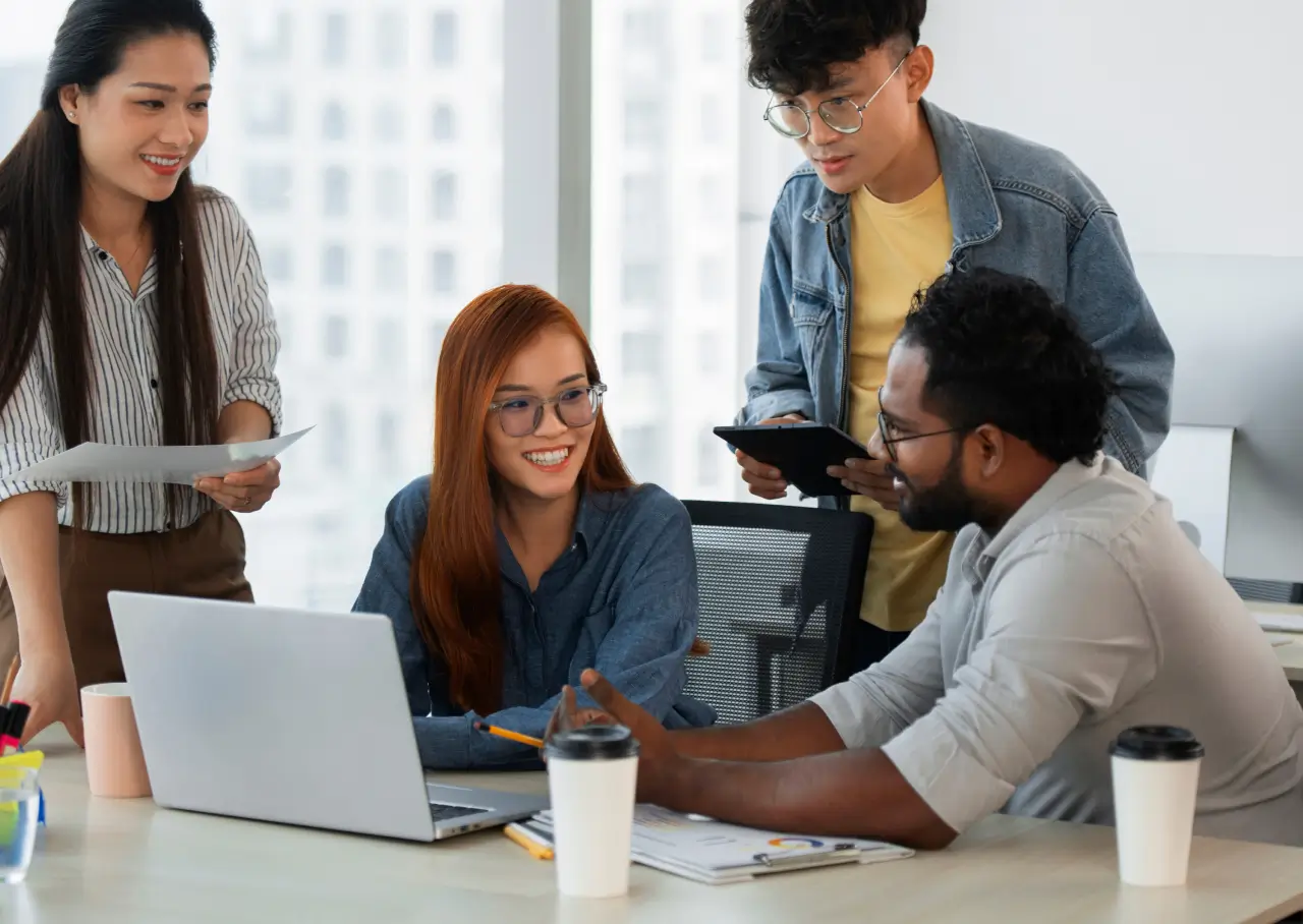 Four colleagues smiling and collaborating around a laptop in a bright office.
