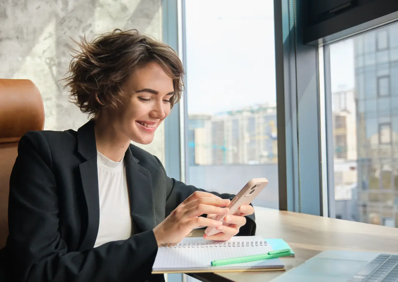 Smiling woman in a black blazer using her smartphone at a desk with notebook and laptop by a window.
