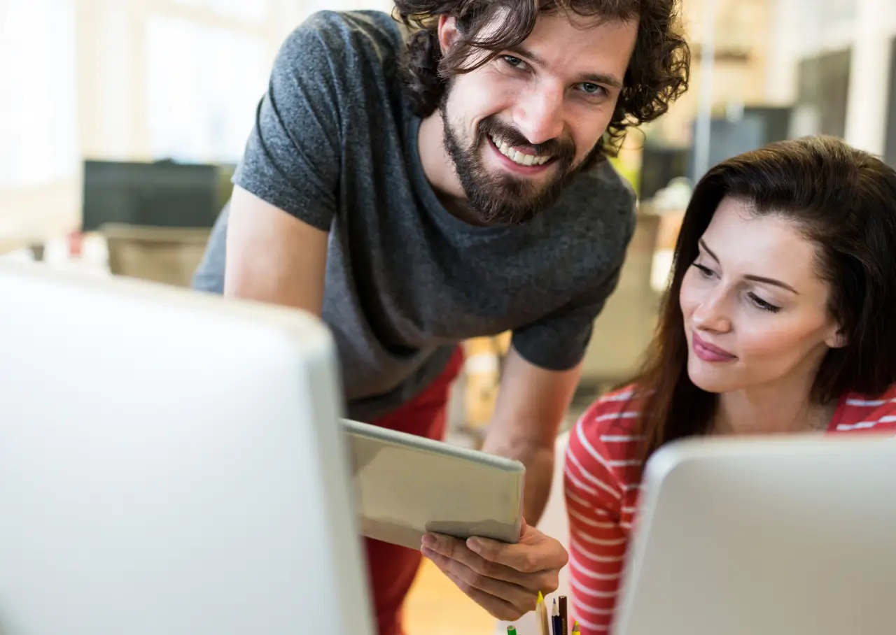 Smiling man holding a tablet and showing something to a woman sitting at a desk with a computer.
