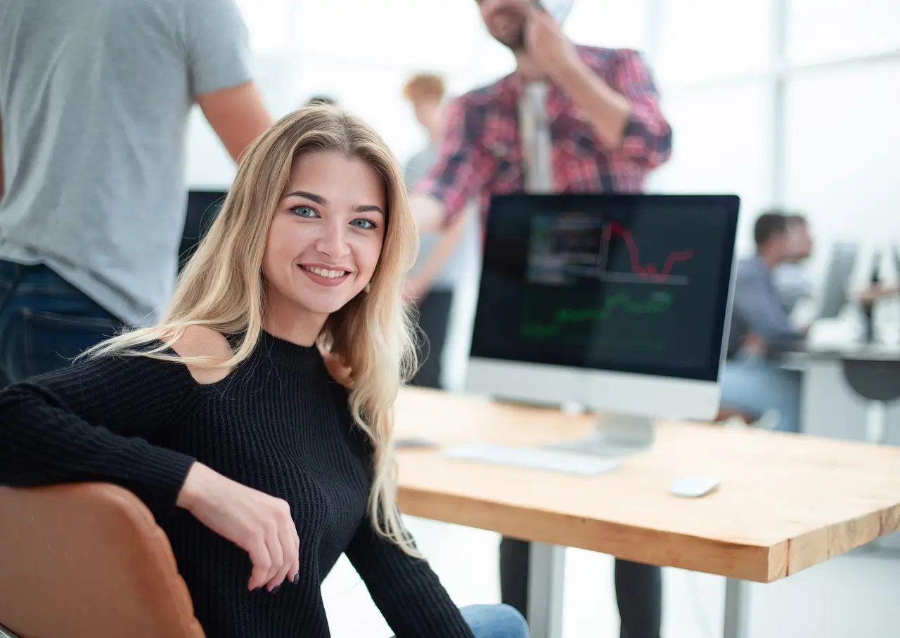 Smiling blonde woman sitting at a desk in an office with a computer monitor displaying graphs in the background.