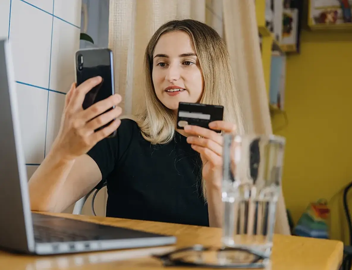 Woman holding a credit card and smartphone, sitting at a table with a laptop, performing Auto Identification with her smartphone before making an online payment.