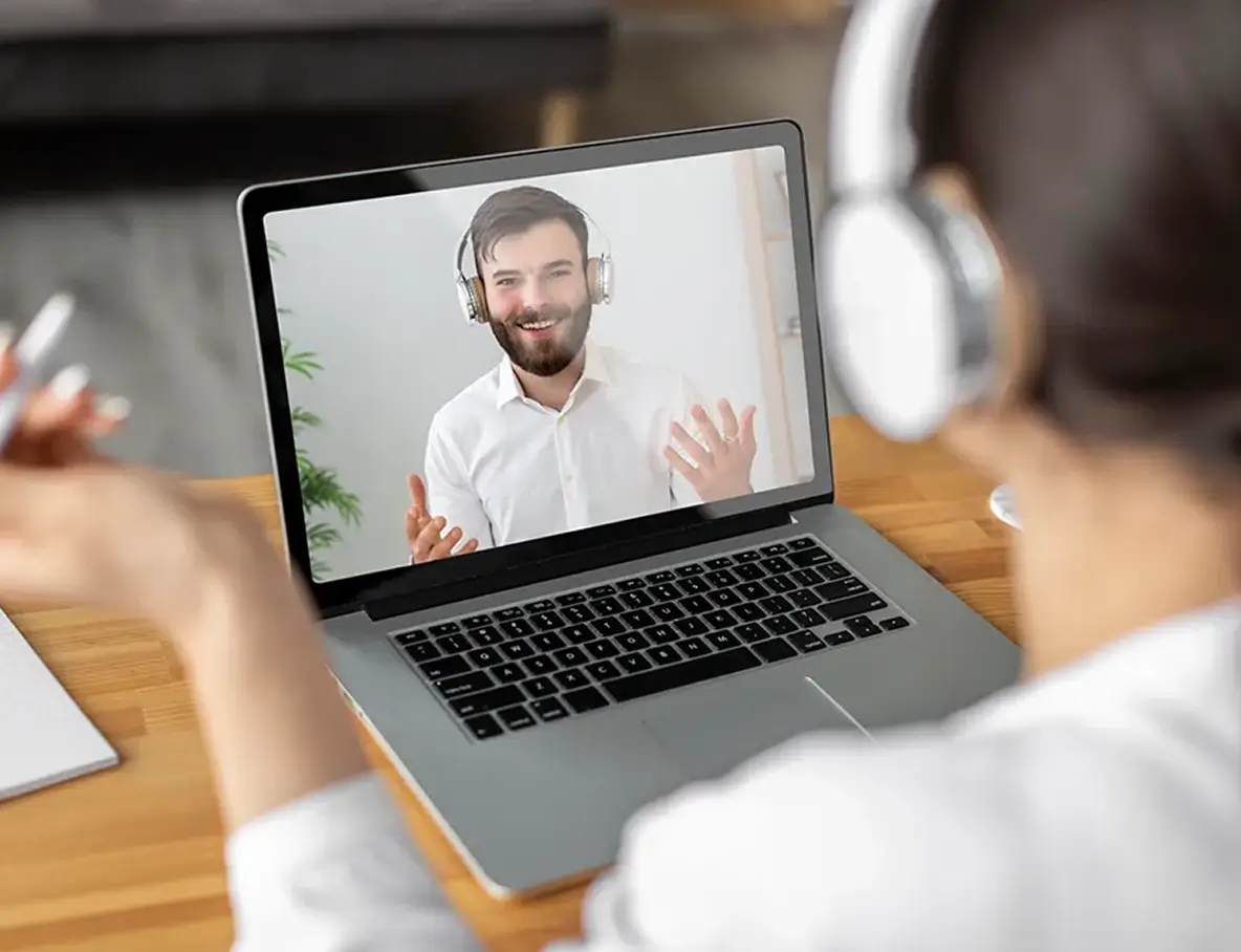 Person wearing headphones attending a video call for video identification, with a smiling agent verifying the persons identity, also wearing headphones visible on the laptop screen.