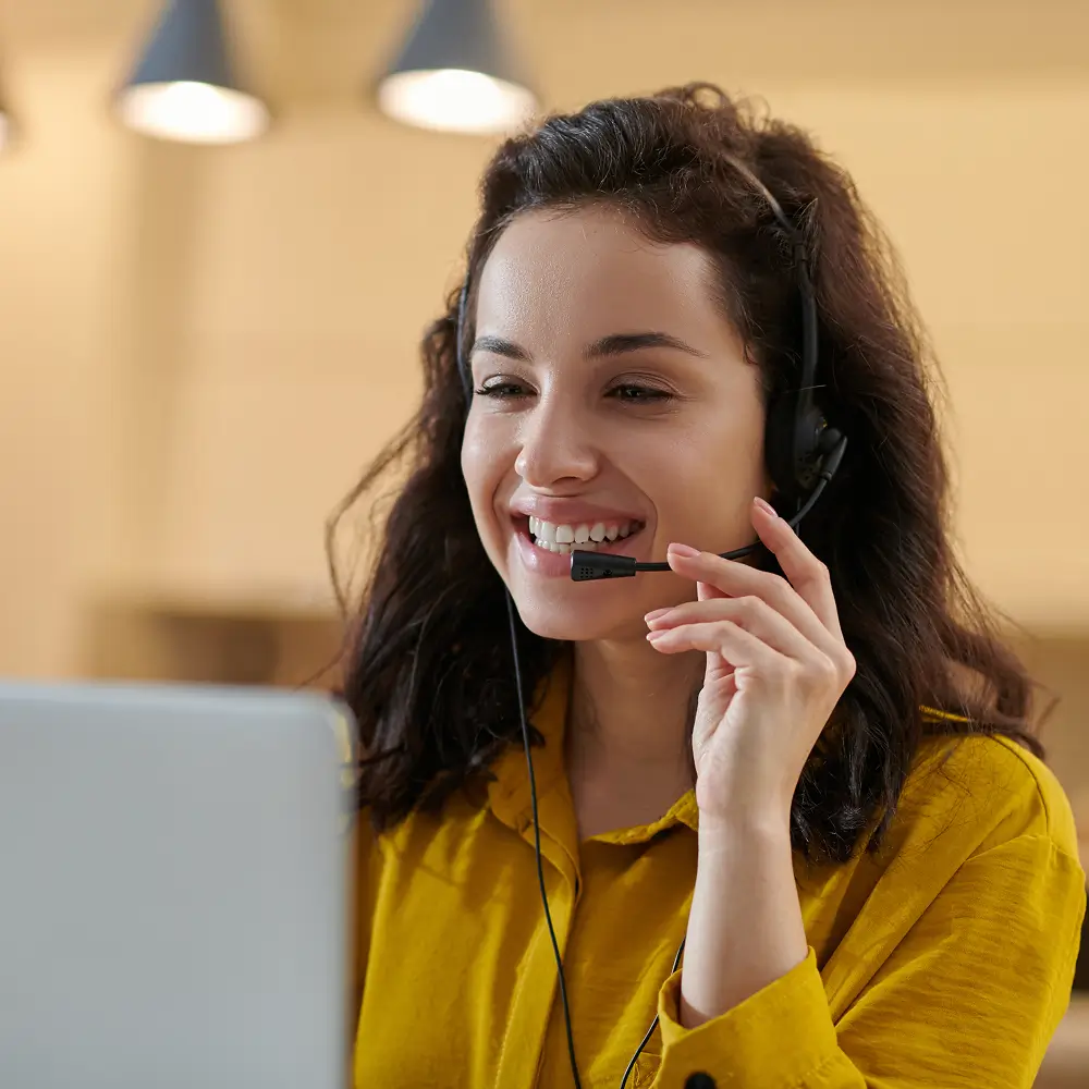 Smiling woman with headset and microphone looking at a laptop, engaged in a video call or customer support.