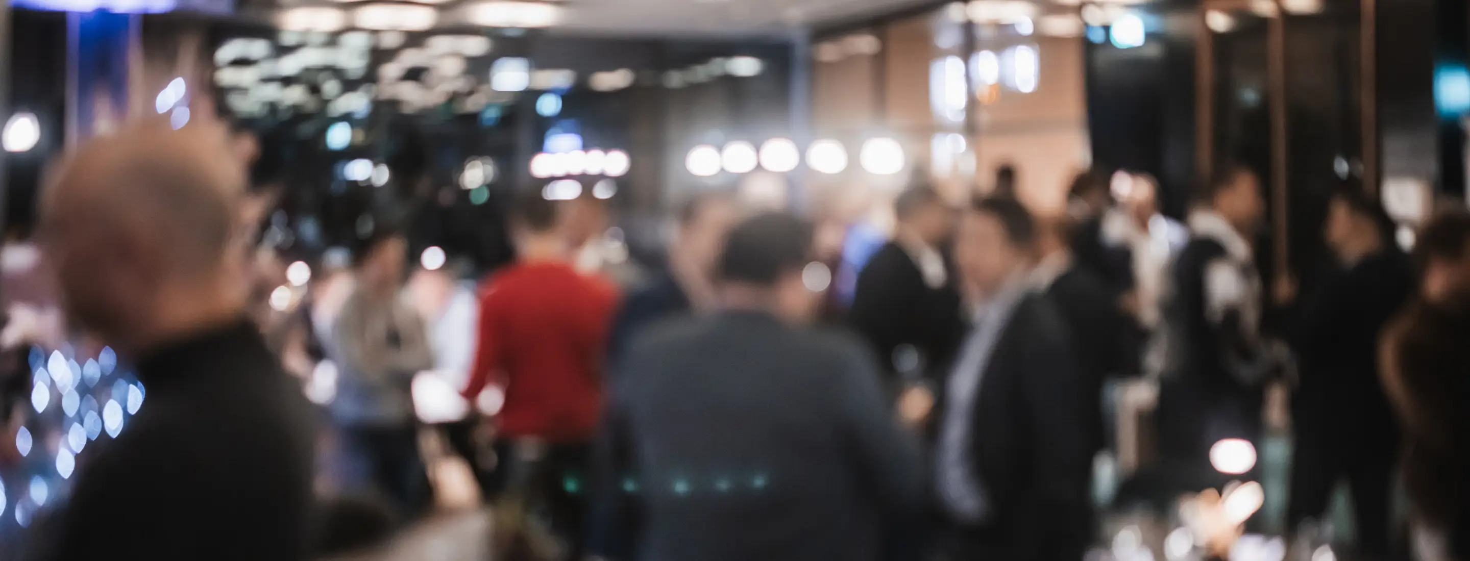 Blurred image of people socializing indoors in a warmly lit room or event space.