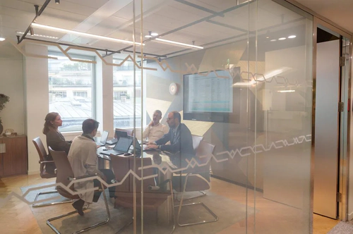 Four people in a meeting in a glass-walled conference room, with laptops and a screen displaying data.