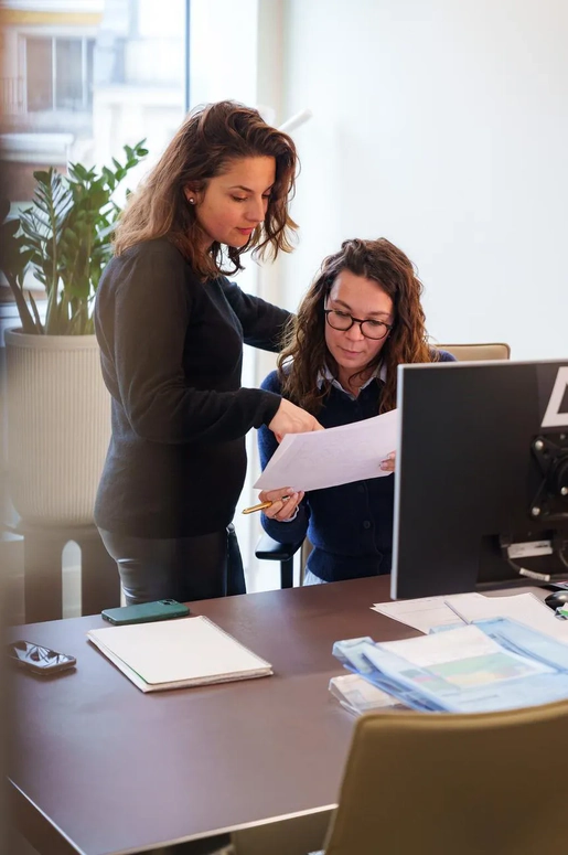 Zwei Frauen sehen sich gemeinsam ein Dokument in einem Büro an, in dem ein Computer und Akten auf dem Schreibtisch liegen.