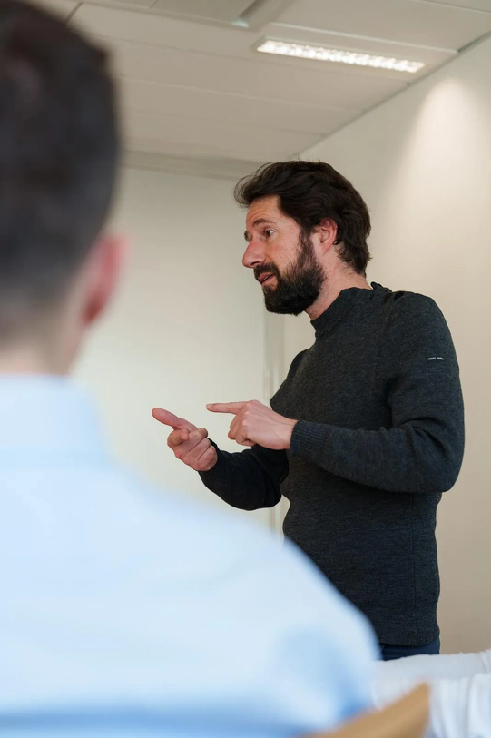 Ein bärtiger Mann in einem dunkelgrauen Pullover, der spricht und mit den Händen in Richtung einer anderen, im Vordergrund unscharfen Person gestikuliert.