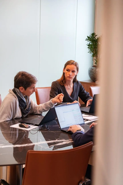 Three people sitting around a black table with laptops, engaged in serious discussion in a modern conference room.