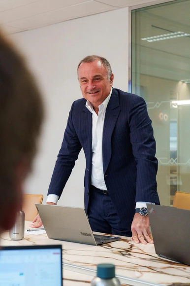 A smiling businessman in a suit standing at a conference table with a laptop open.