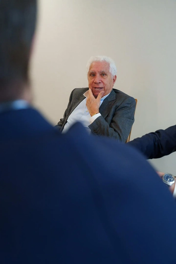 An elderly man with white hair, wearing a reflective gray suit, sitting and talking with two blurry figures in the foreground.