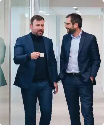 Two men in suits are talking as they walk through an office; one of them is holding a cup of coffee.
