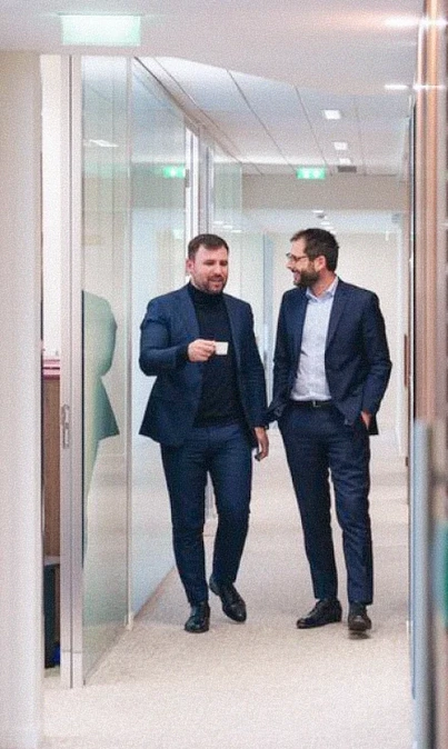 Deux hommes en costume marchant et discutant dans un couloir de bureau moderne.