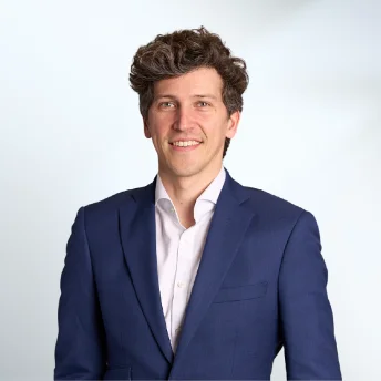 Homme souriant portant un costume bleu et une chemise blanche sur un fond clair.