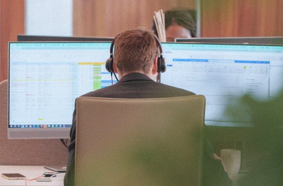 A person seen from behind, wearing headphones, working in front of two large computer monitors displaying charts and data.