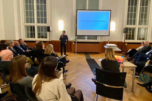 Ein Mann hält einen Vortrag auf einer Veranstaltung, vor ihm sitzt das Publikum, hinter ihm ist ein Bildschirm, auf dem die Frage steht „Welche Herausforderungen sehen Sie?".