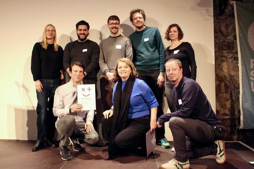 Gruppenfoto von acht Personen, die in zwei Reihen vor einem neutralen Hintergrund stehen. In der vorderen Reihe knien zwei Männer und eine Frau; der Mann links hält eine Urkunde mit vpm Preis für gute Verwaltung. Die hintere Reihe besteht aus fünf Personen, die lächeln und sich locker positionieren. Alle Personen tragen Namensschilder, und die Stimmung wirkt freundlich und entspannt.