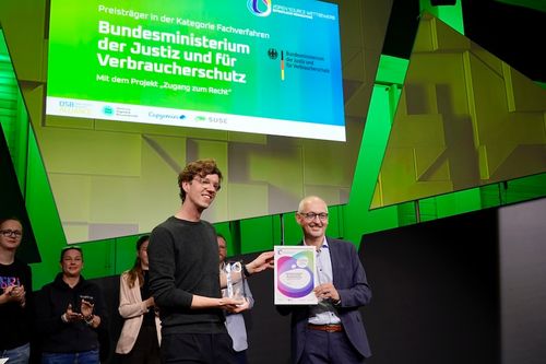 Zwei Männer stehen auf einer Bühne bei der Preisverleihung des Open-Source-Wettbewerbs. Der jüngere Mann links, der einen dunklen Pullover trägt und lächelt, hält eine Trophäe in der Hand. Der ältere Mann rechts, bekleidet mit einem blauen Anzug und einer Brille, hält eine gerahmte Urkunde mit dem Logo des Preises. Im Hintergrund zeigt eine große Leinwand eine Präsentation mit der Aufschrift „Preisträger in der Kategorie Fachverfahren Bundesministerium der Justiz und für Verbraucherschutz Mit dem Projekt 'Zugang zum Recht'“ sowie Logos des Ministeriums und verschiedener Partner des Wettbewerbs.