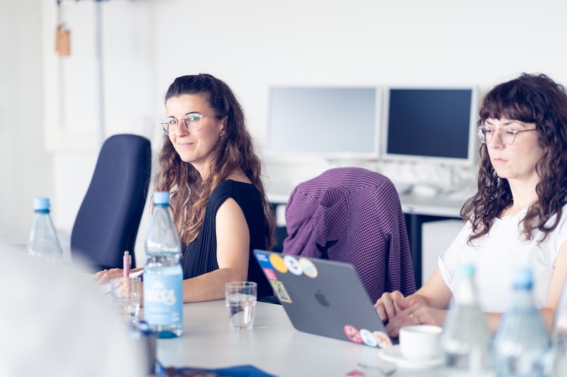 Zwei Frauen sitzen an einem hellen Konferenztisch in einem modernen Büro. Die Frau im Vordergrund links trägt ein schwarzes Oberteil sowie eine Brille und blickt lächelnd in die Kamera. Die Frau rechts daneben arbeitet konzentriert an einem Laptop, der mit verschiedenen Aufklebern verziert ist.