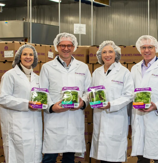 Collaborazione tra Siemens e 80 Acres Farms: quattro rappresentanti in camice bianco che presentano insalate pronte per la spedizione in uno stabilimento di agricoltura verticale.