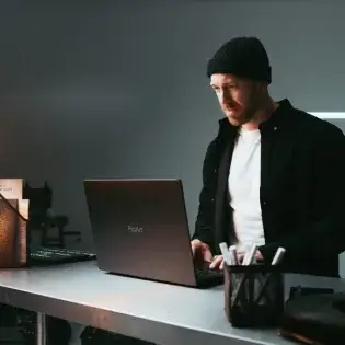 Man in a black beanie and jacket working on a laptop at a desk with office supplies.