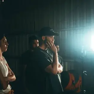 A man in a cap and dark shirt thoughtfully observing a bright light source while others stand nearby in a dimly lit industrial setting.
