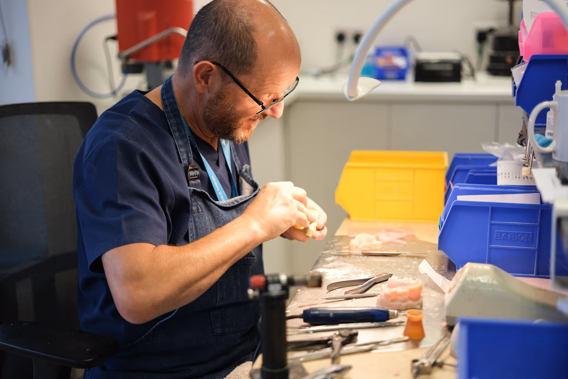 Man wearing glasses and dark blue apron carefully working on a dental mold at a cluttered workstation with dental tools.