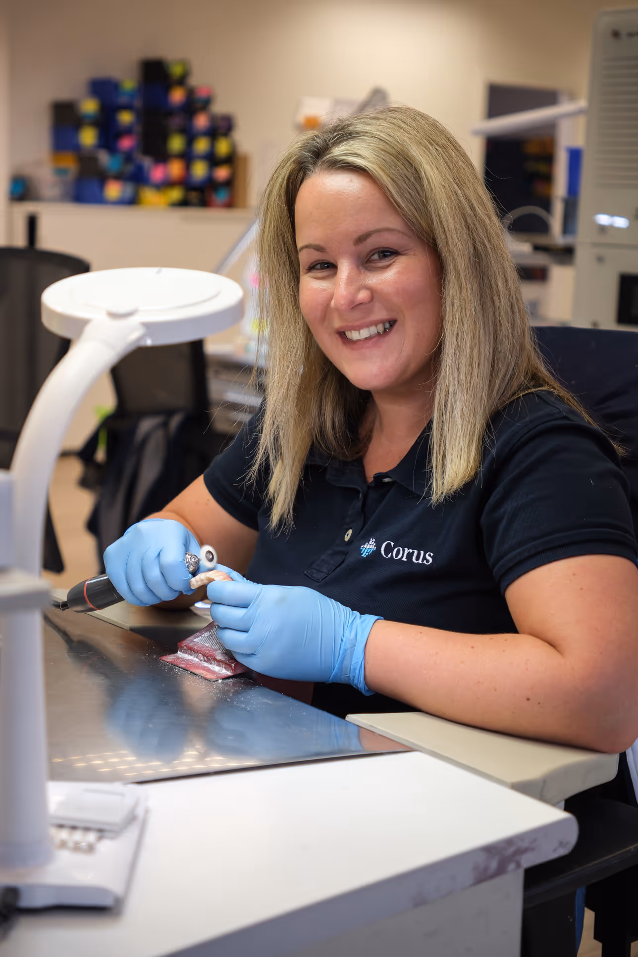 Smiling woman wearing blue gloves working with a rotary tool on a dental mold at a workstation.