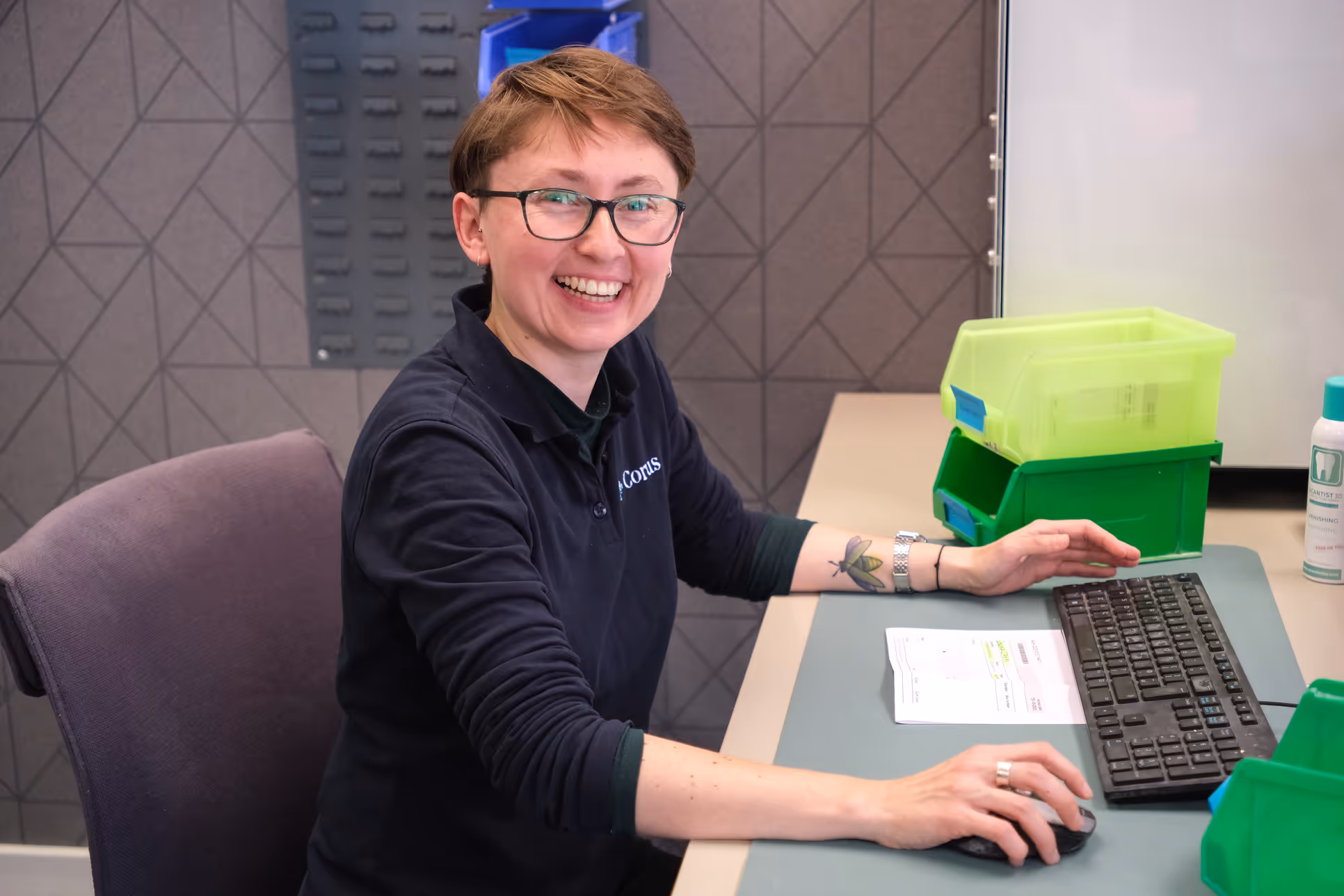 Smiling person with short hair and glasses working on a computer, sitting at a desk with green plastic bins and paperwork.