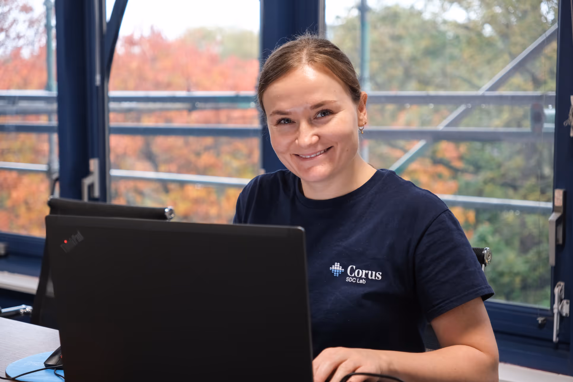 Smiling woman working on a laptop indoors with fall foliage visible through large windows behind her.