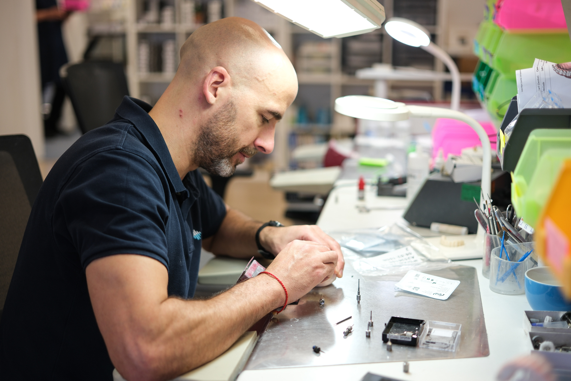 Man assembling small mechanical parts at a well-lit workstation with organized tools and containers.
