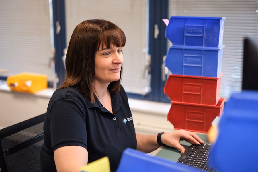Woman with brown hair working on a computer keyboard at a desk with stacked red and blue plastic storage bins in an office setting.