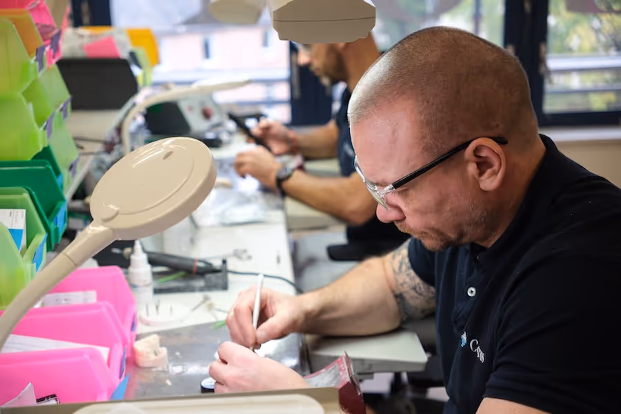 Two men working intently at a workshop bench with tools and colorful storage bins, focusing on detailed manual tasks.