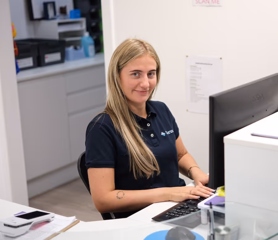 Young woman with long blonde hair wearing a dark polo shirt sitting at a desk, typing on a keyboard in a modern office setting.