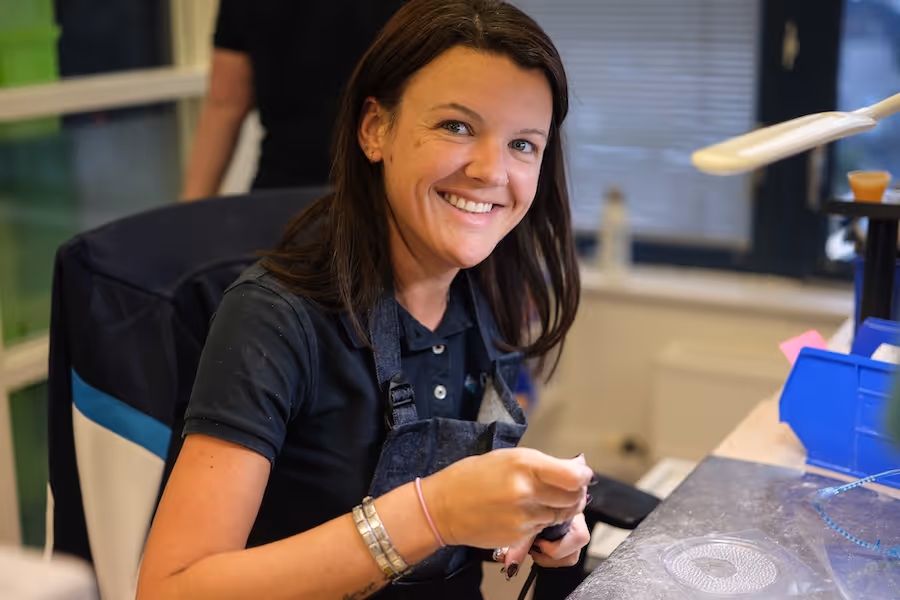 Smiling woman in a black shirt and apron working with jewelry or small craft tools at a well-lit workspace.