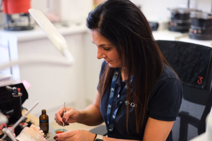 A woman with long dark hair wearing a navy shirt and lanyard is carefully working with tools at a desk in a clinical or lab setting.